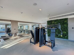 A modern gym with various exercise equipment, a green wall, and a large window. - Chic Studio | Pool View Balcony | Avelon Arjan (Dubai)