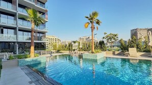 A swimming pool with clear blue water, surrounded by a modern building with balconies and palm trees. - Chic Studio | Pool View Balcony | Avelon Arjan (Dubai)
