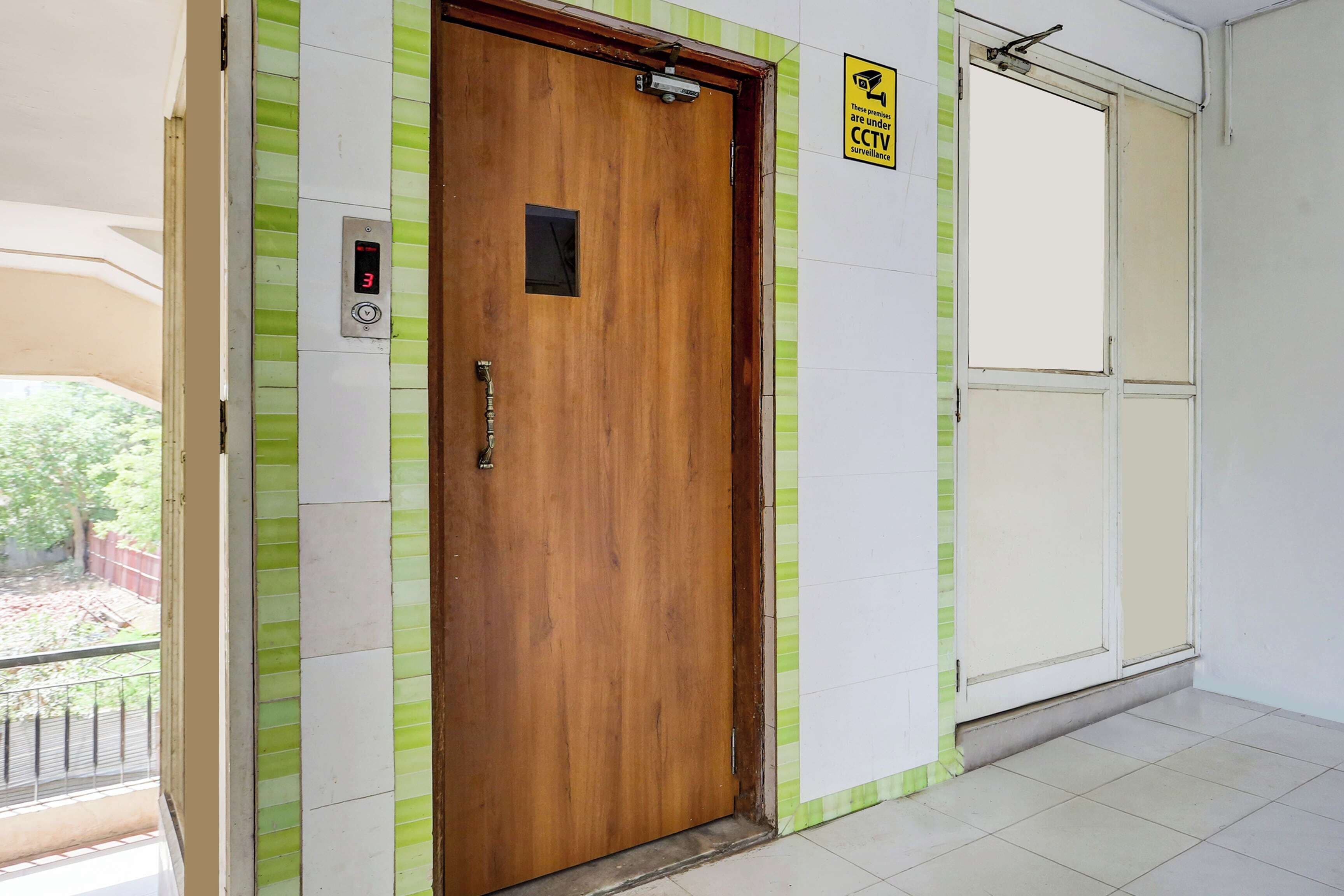 A wooden door with a digital display, a CCTV surveillance sign, and a window with a view of greenery.