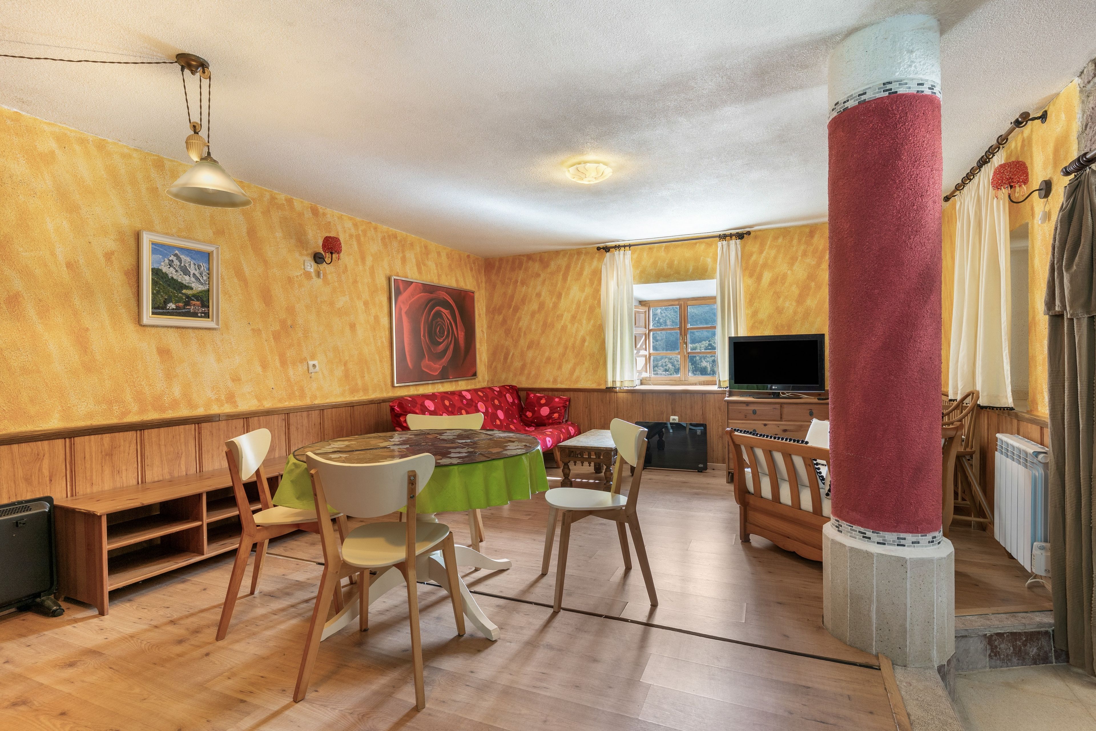 A room with a wooden floor, a round table with chairs, a bench, a television, and a window with curtains.
