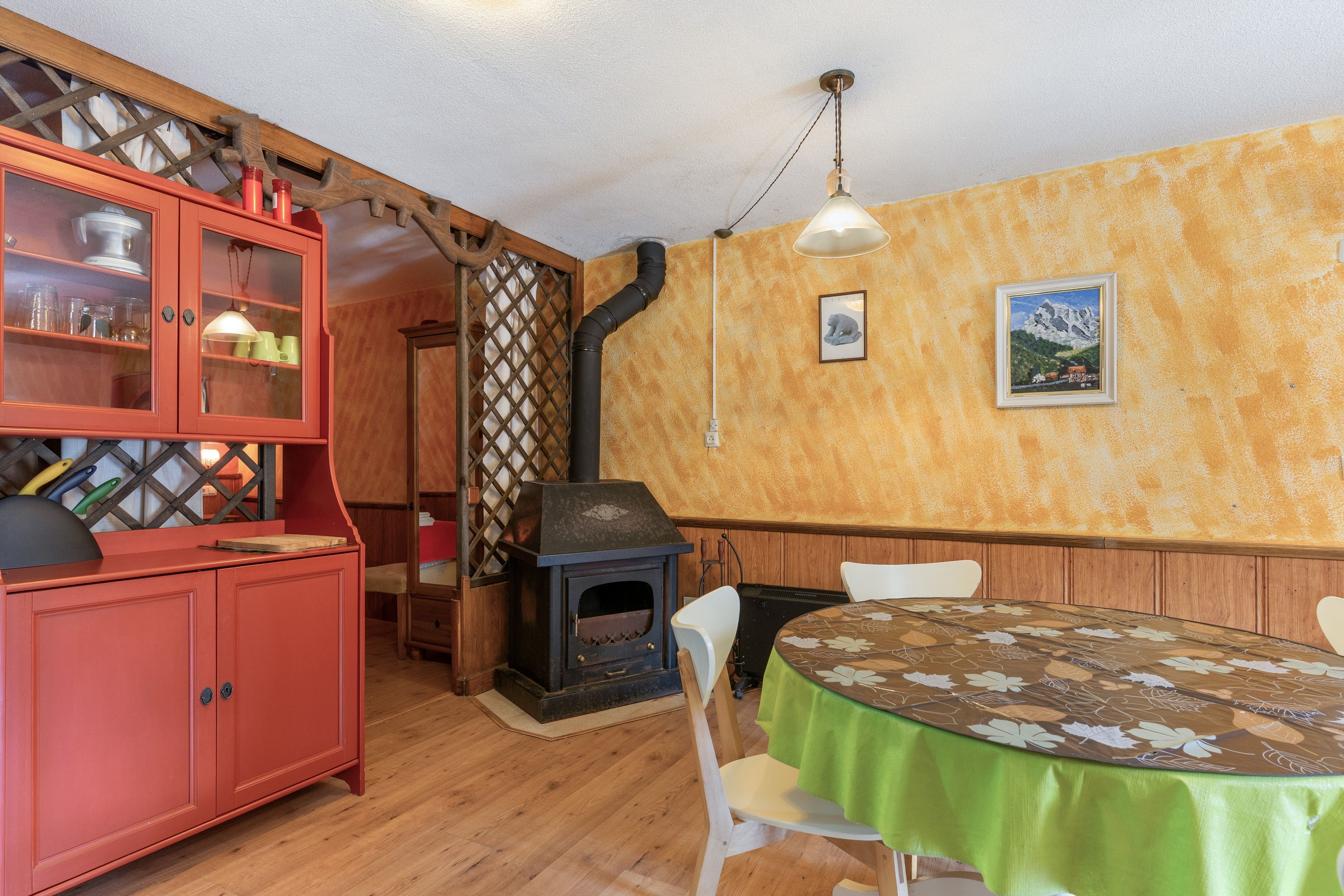 Uma sala de jantar com uma mesa redonda coberta por um pano de mesa verde, cadeiras de madeira, um armário vermelho com portas de vidro e uma lareira a lenha.