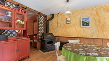 Uma sala de jantar com uma mesa redonda coberta por um pano de mesa verde, cadeiras de madeira, um armário vermelho com portas de vidro e uma lareira a lenha.