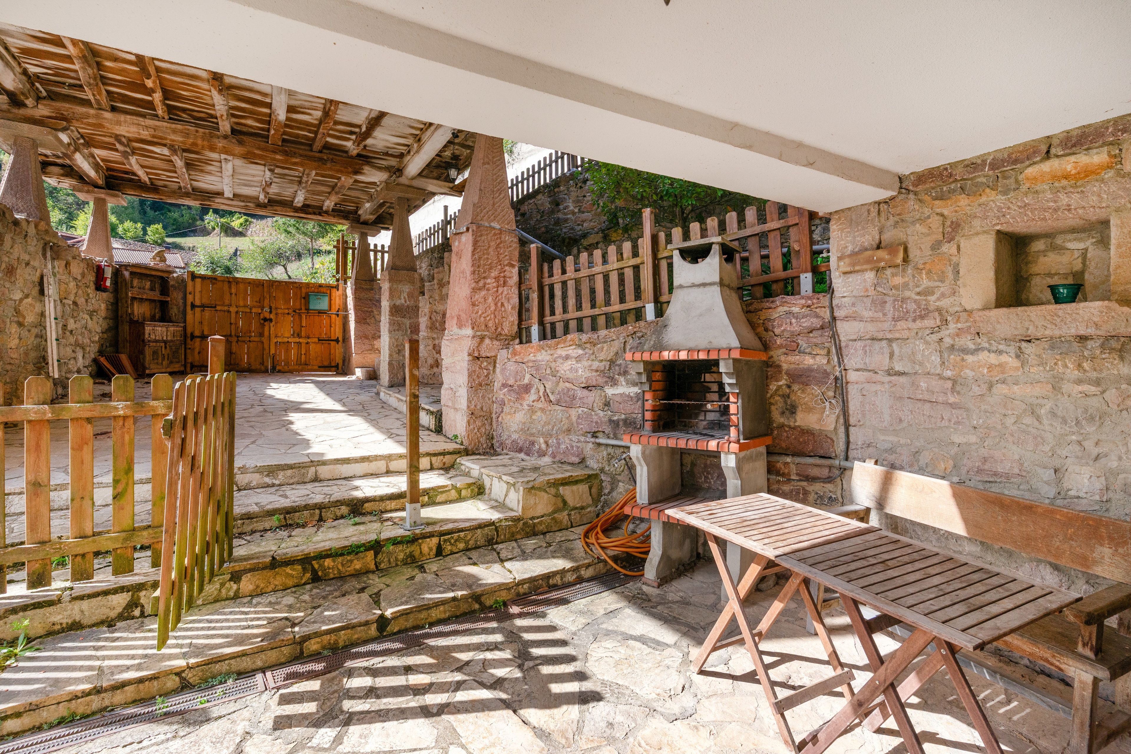 Ein Außenbereich mit Terrasse, einer Steinsäule, einer Holzbank, einem Grill und einer Holztür, die zu einem überdachten Eingang führt.