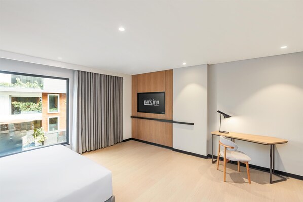 A modern hotel room with a bed, a desk, a chair, a TV, and a view of the outside.