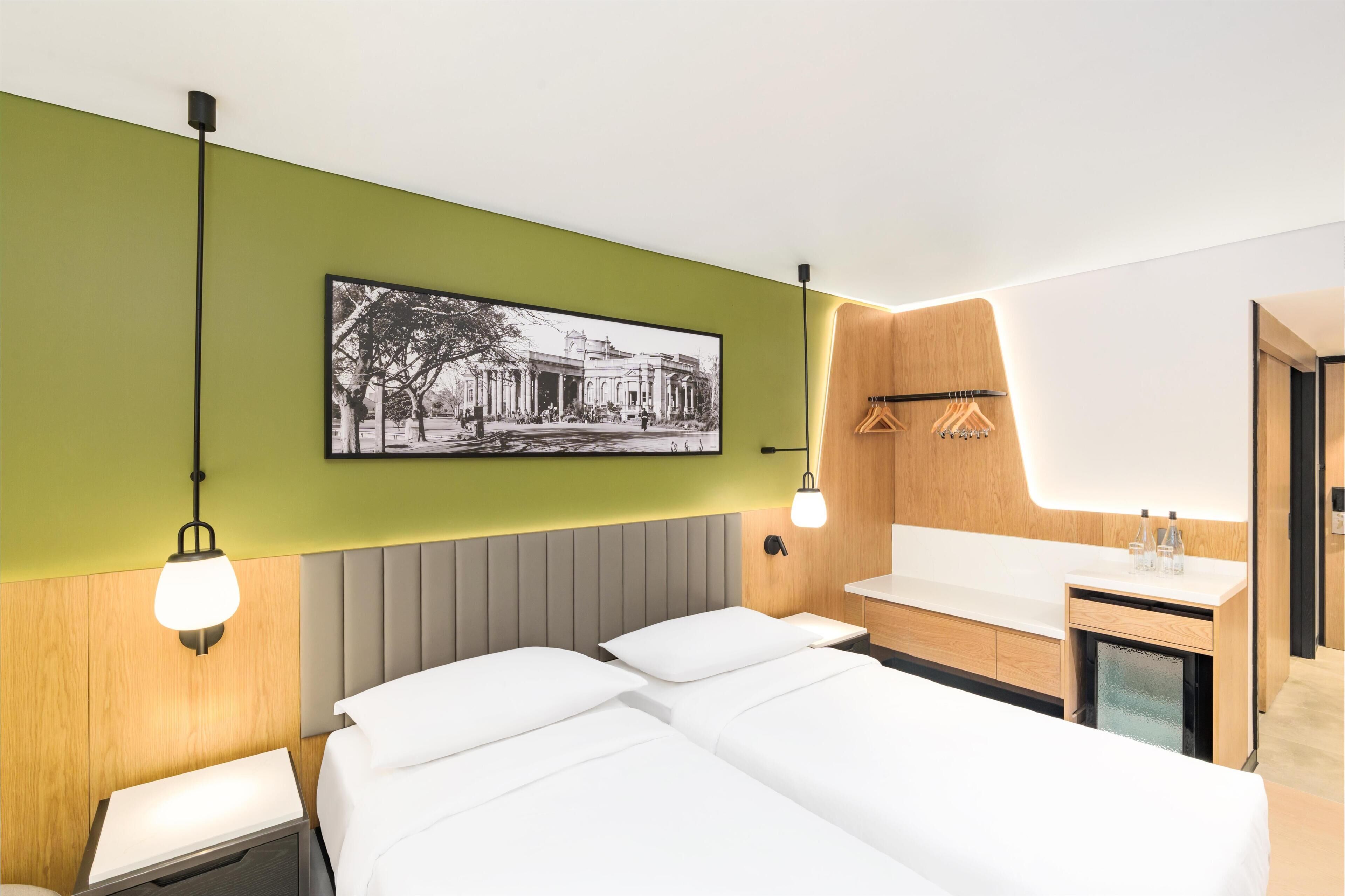 Habitación de hotel con cama, mesitas de noche, un cuadro en la pared y baño con lavamanos y espejo.