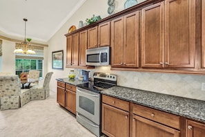 A kitchen with wooden cabinets, a granite countertop, a microwave, a stove, and a dining area with chairs. - Golfing - Matera at Vasari (Bonita Springs)