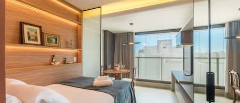 A modern hotel room with a bed, a small dining table with chairs, a balcony with a view, and a TV.
