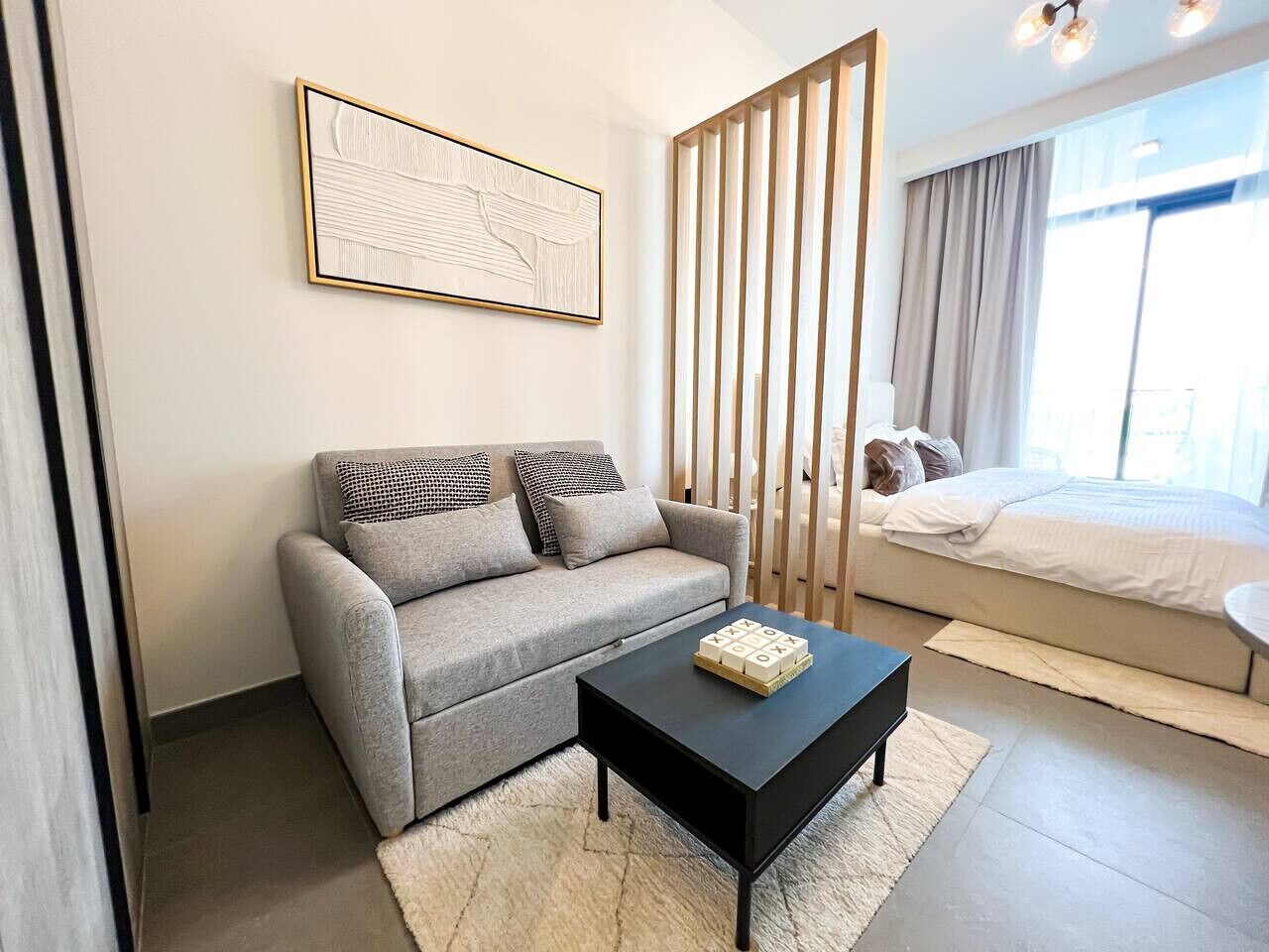 Une chambre moderne avec un canapé gris, une table basse noire et un lit recouvert d’une couette blanche.
