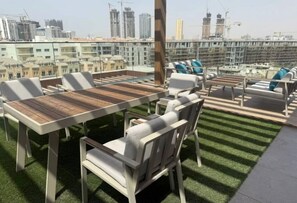 An outdoor seating area with tables and chairs, a grassy surface, and a cityscape view in the background. - Modern Studio in JVC | Pool, Gym & BBQ Access (Dubai)