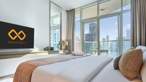 A hotel room with a large bed, a TV mounted on the wall, and a city view through large windows. - Canal View Suite | DAMAC Maison Privé | 4 Guests (Dubai)