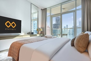 A hotel room with a large bed, a TV mounted on the wall, and a city view through large windows. - Canal View Suite | DAMAC Maison Privé | 4 Guests (Dubai)