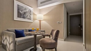 A hotel room with a sofa, a chair, a small table with breakfast, a lamp, and a framed picture on the wall. - Canal View Suite | DAMAC Maison Privé | 4 Guests (Dubai)