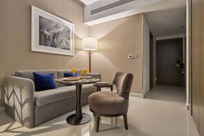 Ein Hotelzimmer mit einer Couch, einem Sessel, einem kleinen Tisch mit Frühstück, einer Lampe und einem gerahmten Bild an der Wand.