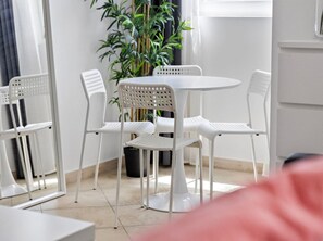 Une pièce avec une table ronde et quatre chaises blanches, une plante en pot et une fenêtre avec des rideaux.