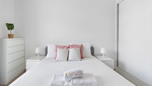 Una cama bien hecha con sábanas blancas y una toalla doblada con la palabra 'WELCOME' escrita en ella. Hay dos mesitas de noche con lámparas, un aparador blanco y una planta en maceta sobre el aparador.