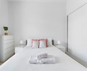 A neatly made bed with white linens and a folded towel with 'WELCOME' written on it. There are two bedside tables with lamps, a white dresser, and a potted plant on the dresser. - Cozy City Studio for 4 Guests - Regent House 2 (Dubai)
