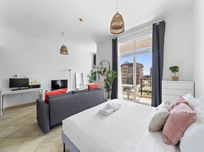 A modern hotel room with a large bed, a sofa, a TV, and a balcony with a view of the city. - Cozy City Studio for 4 Guests - Regent House 2 (Dubai)