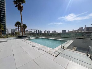 A rooftop swimming pool with a glass railing, surrounded by palm trees and modern buildings in the background. - Chic & Cozy Studio in JVC | Top Amenities & Views! (Dubai)