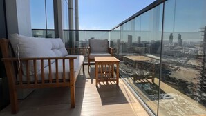 En balkon med træbænk og stol, et lille bord og udsigt over byen gennem store glasvinduer.