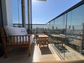 A balcony with a wooden bench and chair, a small table, and a city view through large glass windows. - Chic & Cozy Studio in JVC | Top Amenities & Views! (Dubai)