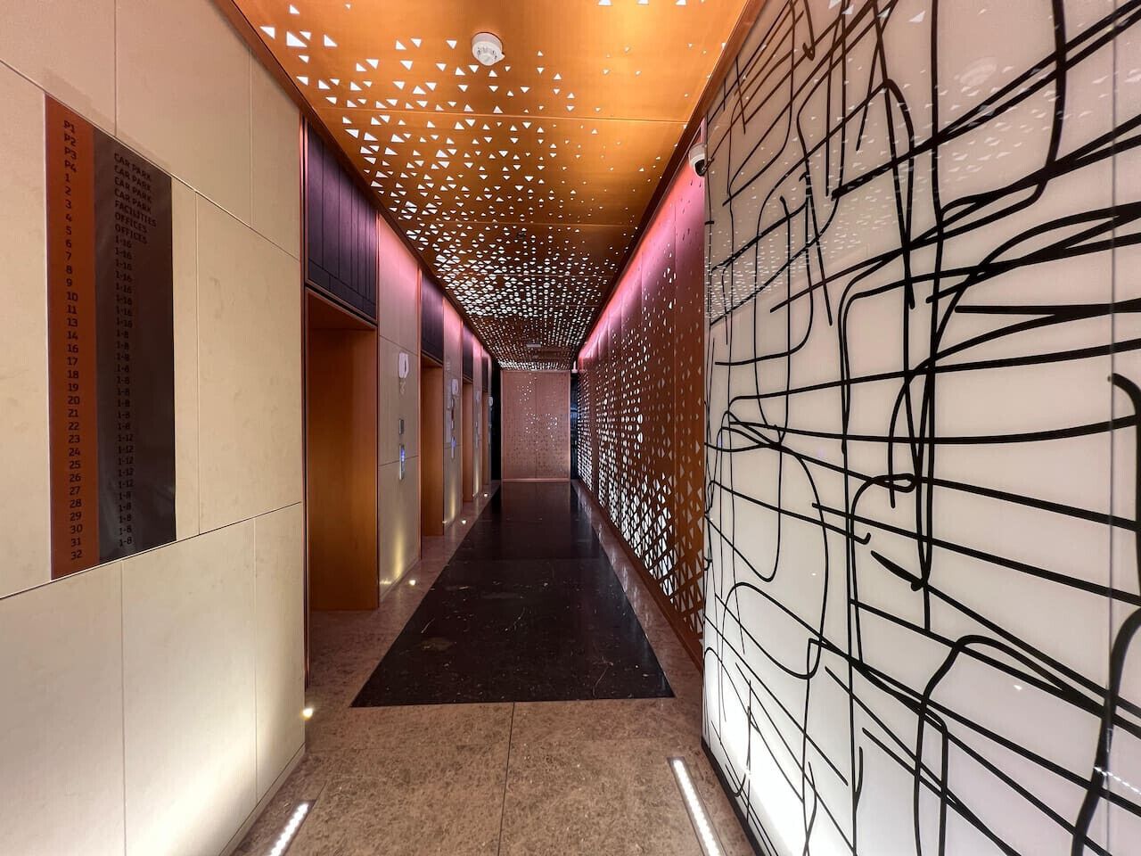 Un couloir recouvert d’un tapis noir, un mur orné d’un motif géométrique et un plafond percé de découpes triangulaires.