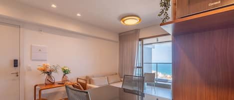 A modern hotel room with a dining area, a sofa, a large window with a view, and a wooden cabinet.