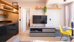 A modern living room with a flat-screen TV, a dining area with yellow chairs, a kitchen with a black countertop, and a hallway leading to a door. - Apartment w/ sea View in Building w/ Pool Edf1303 (Salvador)