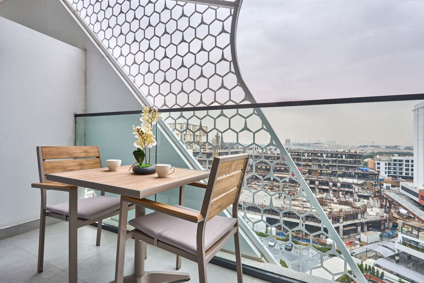 A balcony with a wooden table, two chairs, a vase with flowers, and a view of a cityscape with buildings and construction.