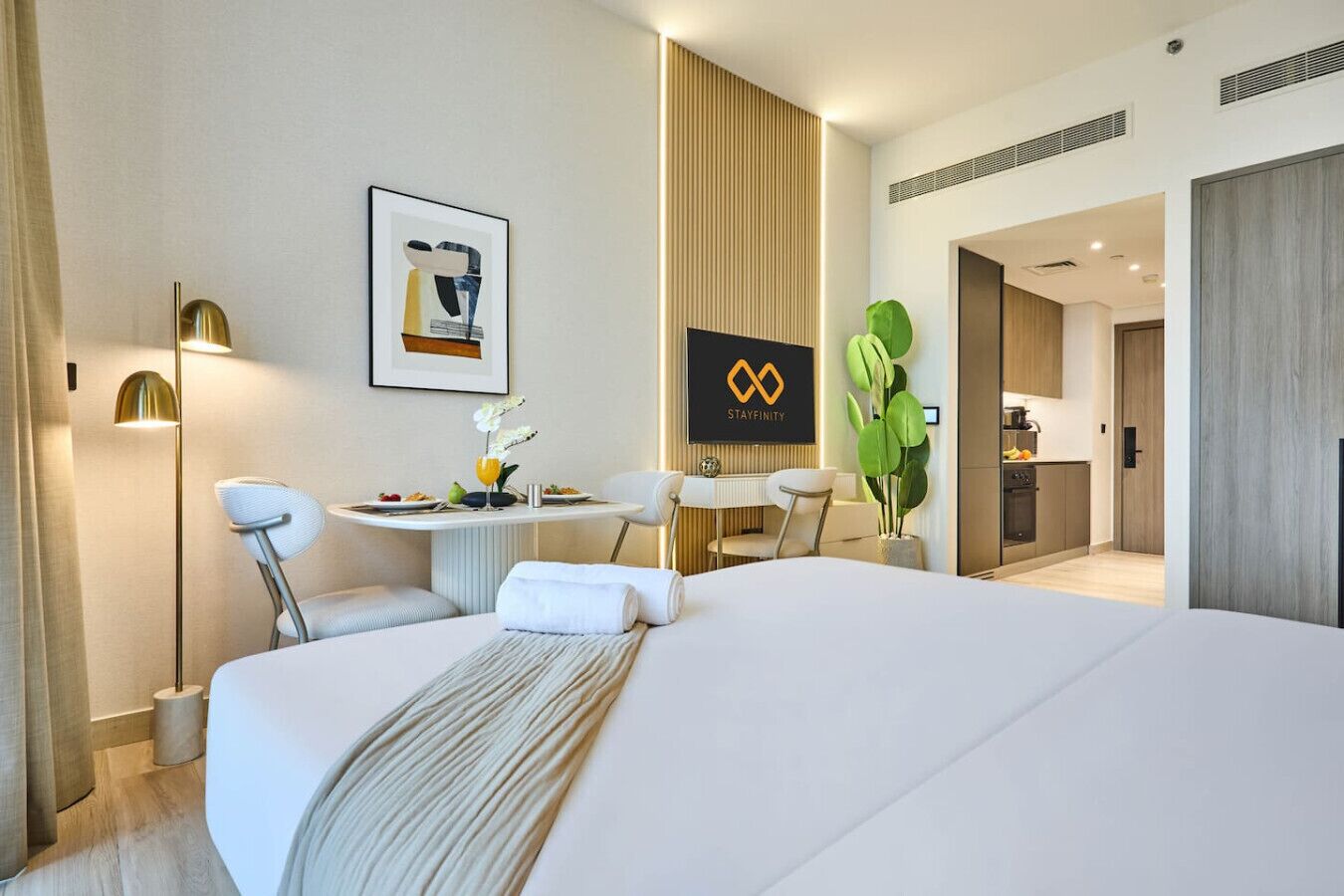 Quarto de hotel com uma cama grande, uma mesa de jantar com cadeiras, uma pequena cozinha e uma obra de arte na parede.