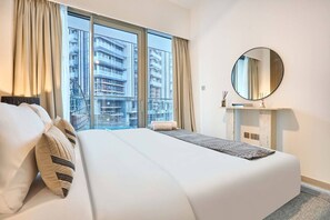 Ein Hotelzimmer mit einem großen Bett, einem runden Spiegel, einem kleinen Konsolentisch und Blick auf eine Skyline durch raumhohe Fenster.