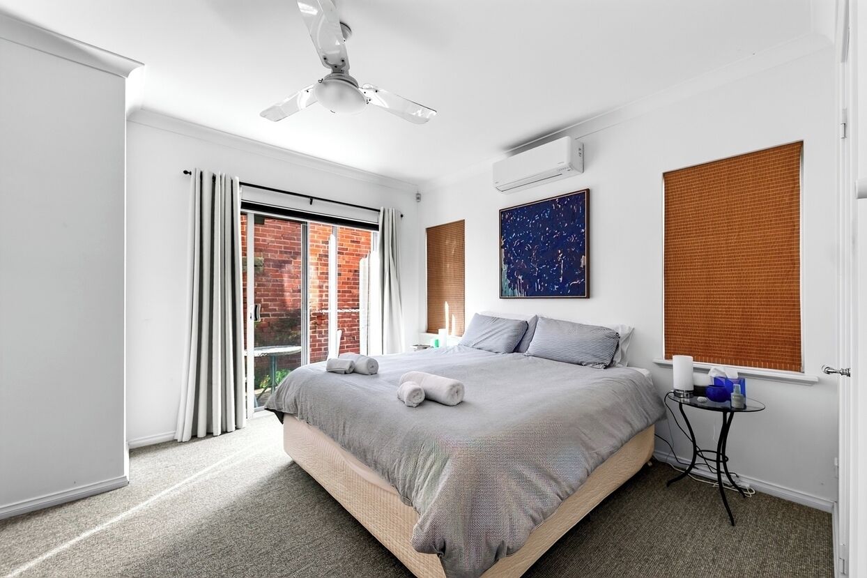 A modern bedroom with a large bed, a ceiling fan, a window with blinds, and a small bedside table.