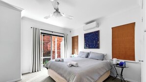 A modern bedroom with a large bed, a ceiling fan, a window with blinds, and a small bedside table. - Two-Storey Cottesloe Garden Retreat with Study & Lavender Charm (Cottesloe)