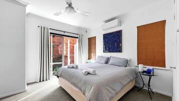 A modern bedroom with a large bed, a ceiling fan, a window with blinds, and a small bedside table.