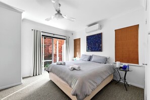 Quarto moderno com cama grande, ventilador de teto, janela com persianas e mesinha de cabeceira.