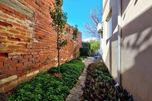 Un camí estret delimitat per una paret de maó i un edifici blanc, amb un arbre petit i plantes al llarg del camí.