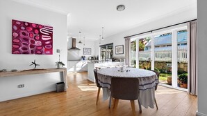A modern kitchen with white cabinets, a dining area with a round table, and a large window with a view of a white fence and greenery. - Two-Storey Cottesloe Garden Retreat with Study & Lavender Charm (Cottesloe)
