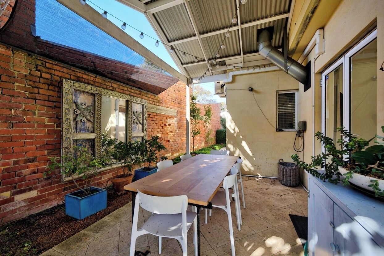 An outdoor patio with a wooden table and white chairs, surrounded by potted plants and a brick wall with decorative elements.
