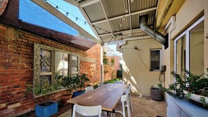 An outdoor patio with a wooden table and white chairs, surrounded by potted plants and a brick wall with decorative elements. - Two-Storey Cottesloe Garden Retreat with Study & Lavender Charm (Cottesloe)