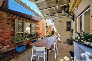 Um pátio externo com mesa de madeira e cadeiras brancas, rodeado por plantas em vasos e uma parede de tijolos com detalhes decorativos.