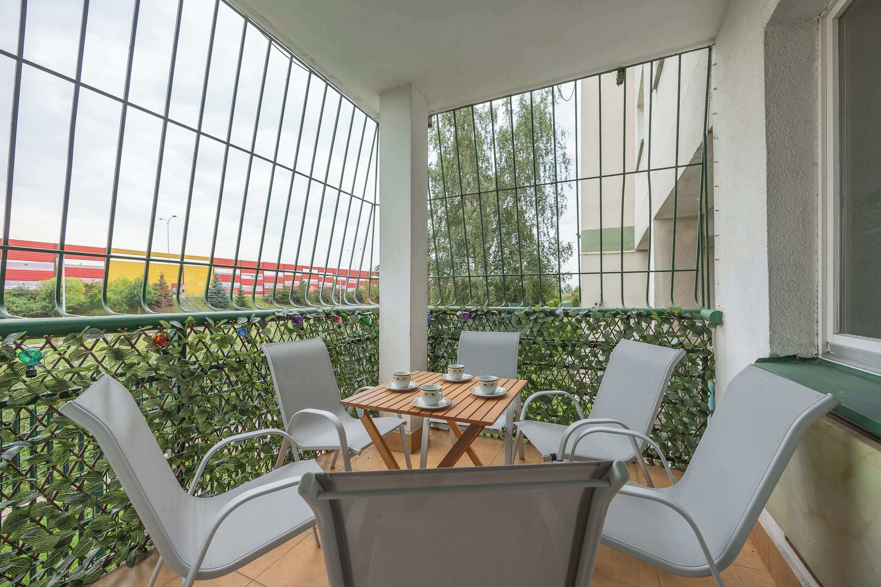 Een balkon met een tafel en stoelen, uitzicht op een gebouw met rode en gele strepen, en een groene hekken.