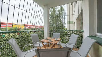 Een balkon met een tafel en stoelen, uitzicht op een gebouw met rode en gele strepen, en een groene hekken.