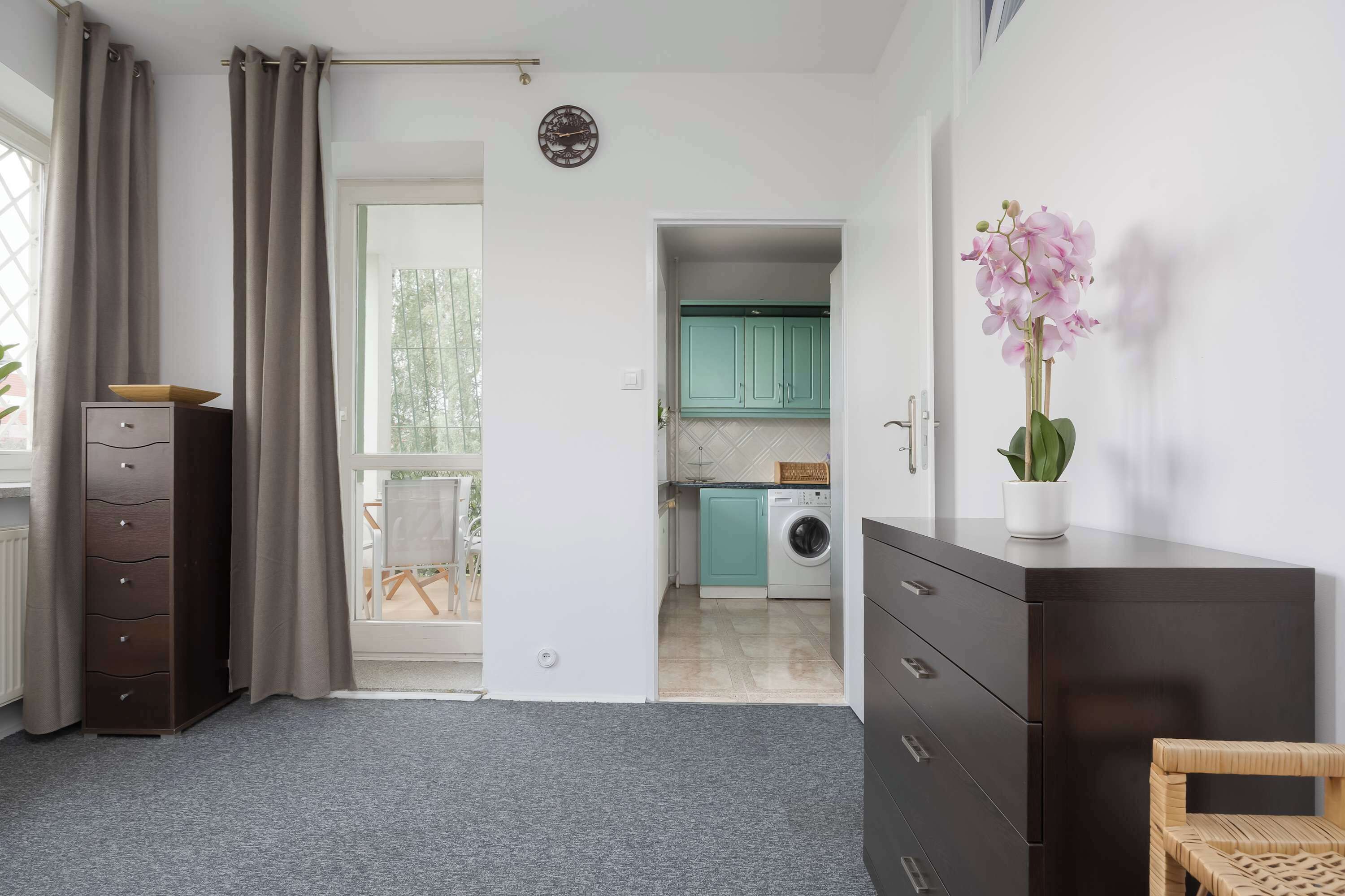 Een modern appartement met een grijs vloerkleed, een houten kast, een witte inbouwkast, een eethoek met een tafel en stoelen, en een keuken met blauwgroene kastjes en een wasmachine.