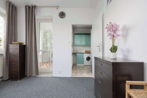 Eine moderne Wohnung mit grauem Teppich, einem Holzschrank, einem weißen Kleiderschrank, einem Essbereich mit Tisch und Stühlen sowie einer Küche mit türkisblauen Schränken und einer Waschmaschine.