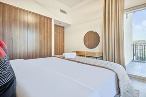 A hotel room with a large bed, a wooden wardrobe, a round mirror, a small bedside table, and a balcony with a view. - Family-Friendly 2BR in Remraam with Pool View (Dubai)