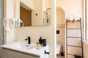 Ein Badezimmer mit weißem Waschbecken, einem schwarzen Wasserhahn und einem Spiegel darüber. Auf dem Waschbecken liegen Toilettenartikel und ein Handtuchhalter mit Handtüchern.