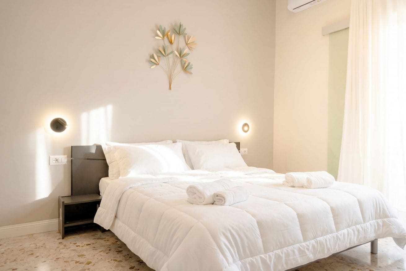 Una cama bien hecha con ropa blanca y dos almohadas, una mesita de noche con una lámpara, y una lámpara de pared.