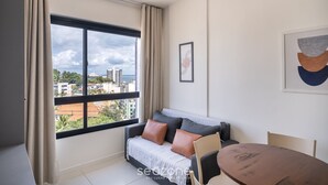 A modern living room with a large window, a sofa, a dining table, and a framed abstract painting. - Apartment 400m From Barra Beach Blb1306 (Salvador)