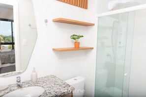 Ein Badezimmer mit einem weißen Waschbecken, einer Toilette und einer Duschkabine aus Glas. Über dem Waschbecken befinden sich Holzregale und auf einem der Regale steht ein Blumentopf.