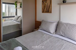 Ein Schlafzimmer mit großem Fenster, einem Bett mit gemusterten Betttextilien, einem kleinen Nachttisch und einer Wandpflanze.