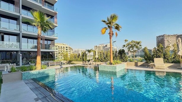 A swimming pool with clear blue water, surrounded by palm trees and modern buildings. - Chic Studio | Pool View Balcony | Avelon Arjan (Dubai)